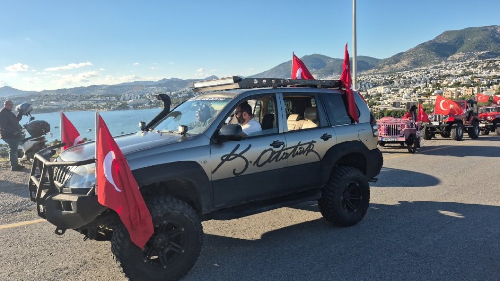 Bir grup araç, Türk bayraklarıyla süslenmiş şekilde bir yolda ilerliyor. Ön planda, içinde sürücü ve yolcunun bulunduğu siyah bir SUV araç yer alıyor. Arka planda, deniz manzarası ve dağlık bir alan görünmekte.