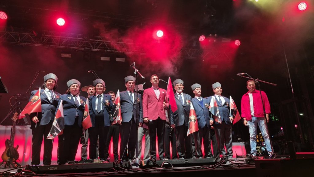Bir müzik etkinliğinde, farklı kostümler giymiş ve bayraklar tutan bir grup erkek sanatçı sahnede duruyor.