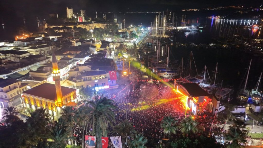 Gece manzarasında tarihi kalenin yanı başında kalabalık bir etkinlik alanı, canlı sahne ve limanda demirli yatlar.