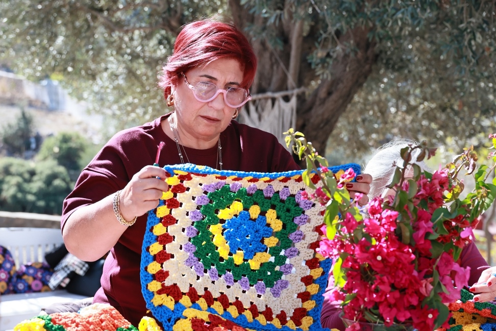 Renkli bir haşiye örneği elinde tutan kadın, arka planda yeşil ağaçlar ve çiçekler ile birlikte görünmektedir.